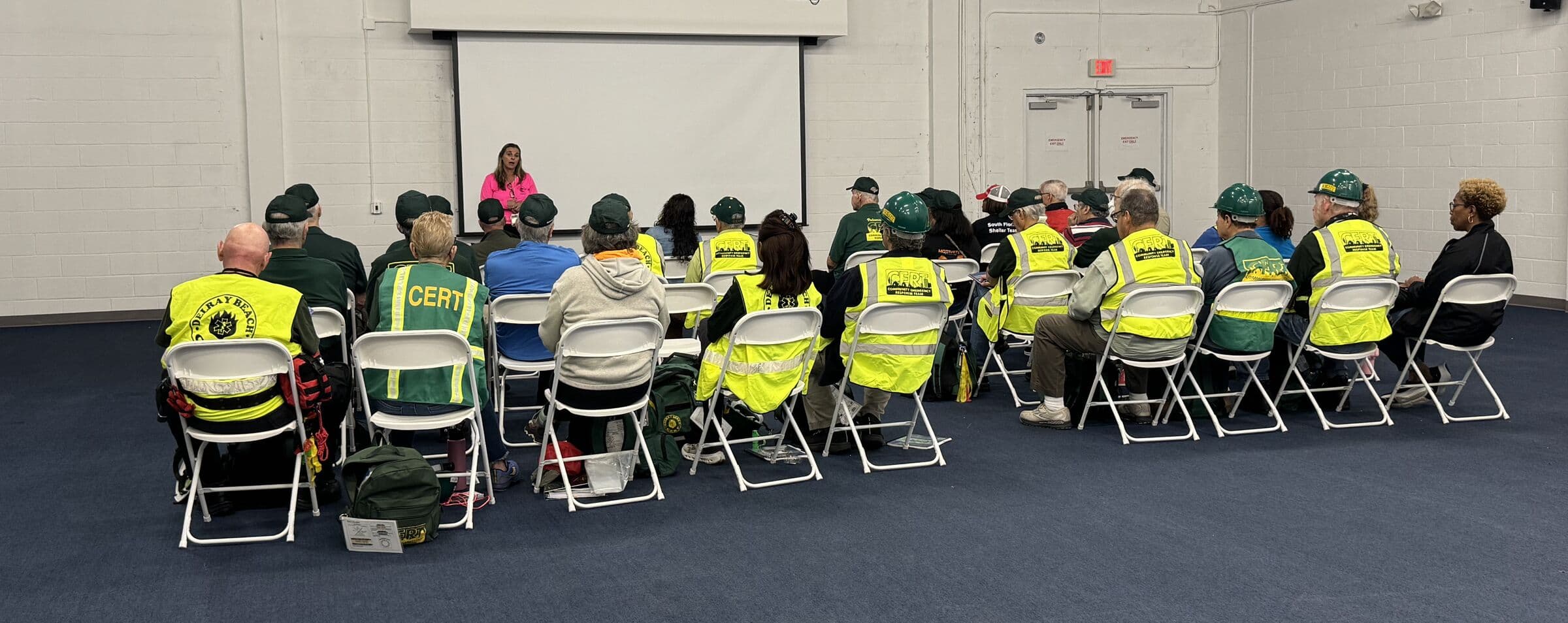 About two dozen people in high-visibility CERT vests seated in rows of folding chairs in a plain block-walled room, facing a presenter standing at a projection screen