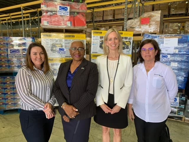 Four people standing together in a warehouse aisle in front of pallets of stacked boxes