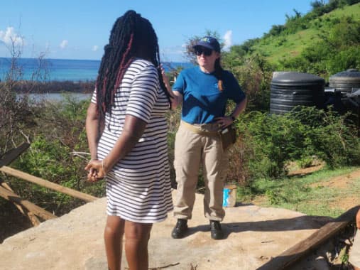 Two people talking on a concrete slab on a hillside, with water tanks behind them and the ocean in the distance