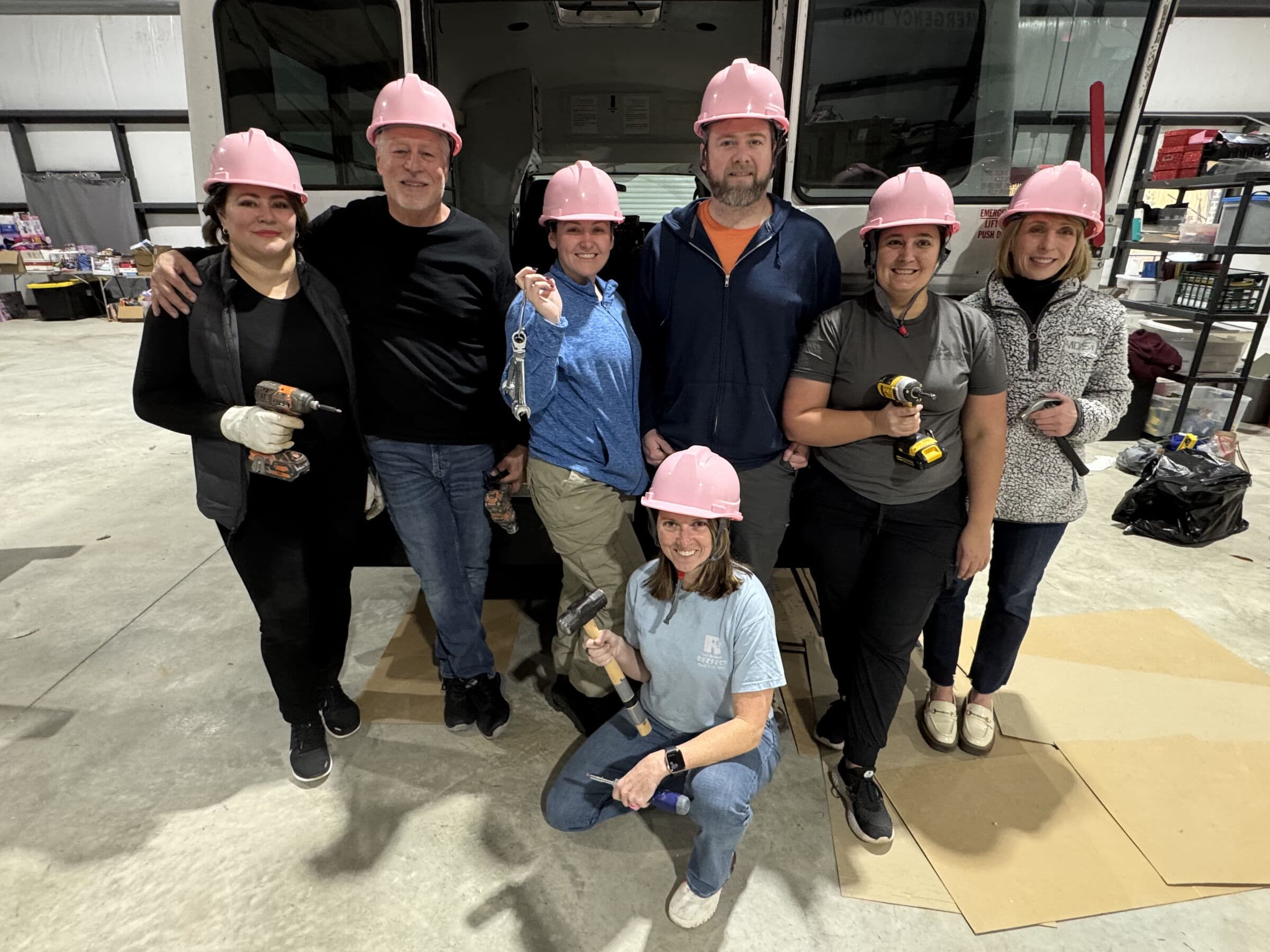 Seven people wearing pink hard hats posed together in front of the open rear doors of a box truck inside a warehouse, several holding cordless drills and hand tools