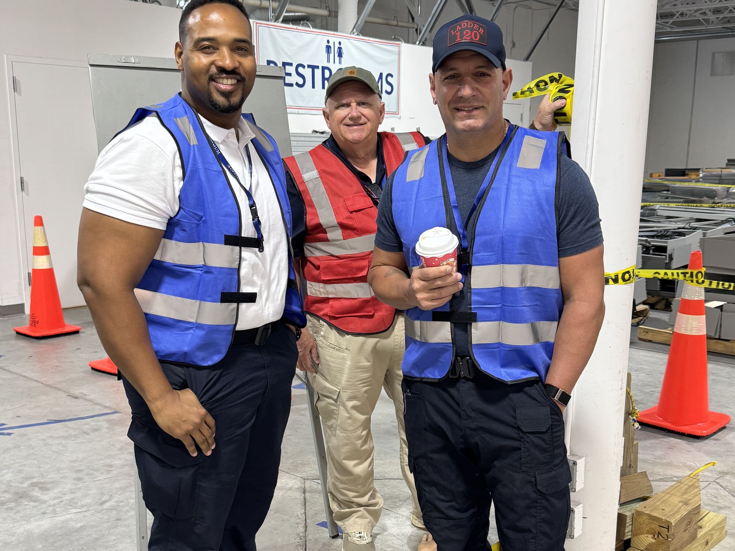 Three men in high-visibility vests standing together in a warehouse bay, one holding a paper cup, with orange traffic cones and yellow caution tape behind them