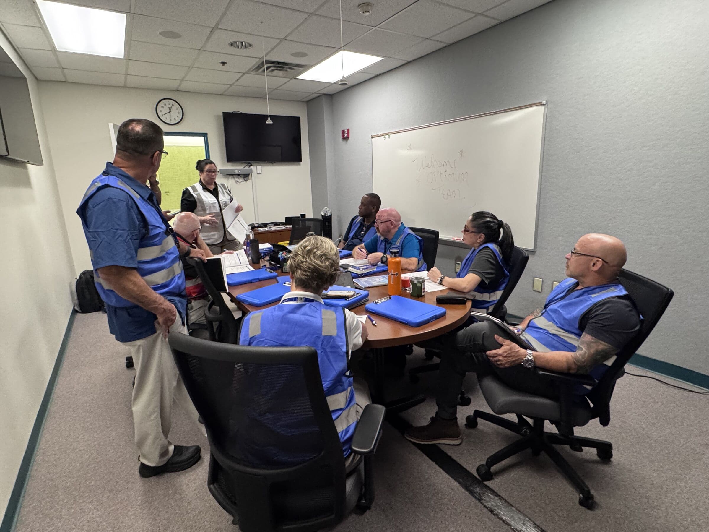 Eight people in blue high-visibility vests around an oval meeting table, one standing with papers in hand addressing the others, a wall clock and a handwritten whiteboard behind them