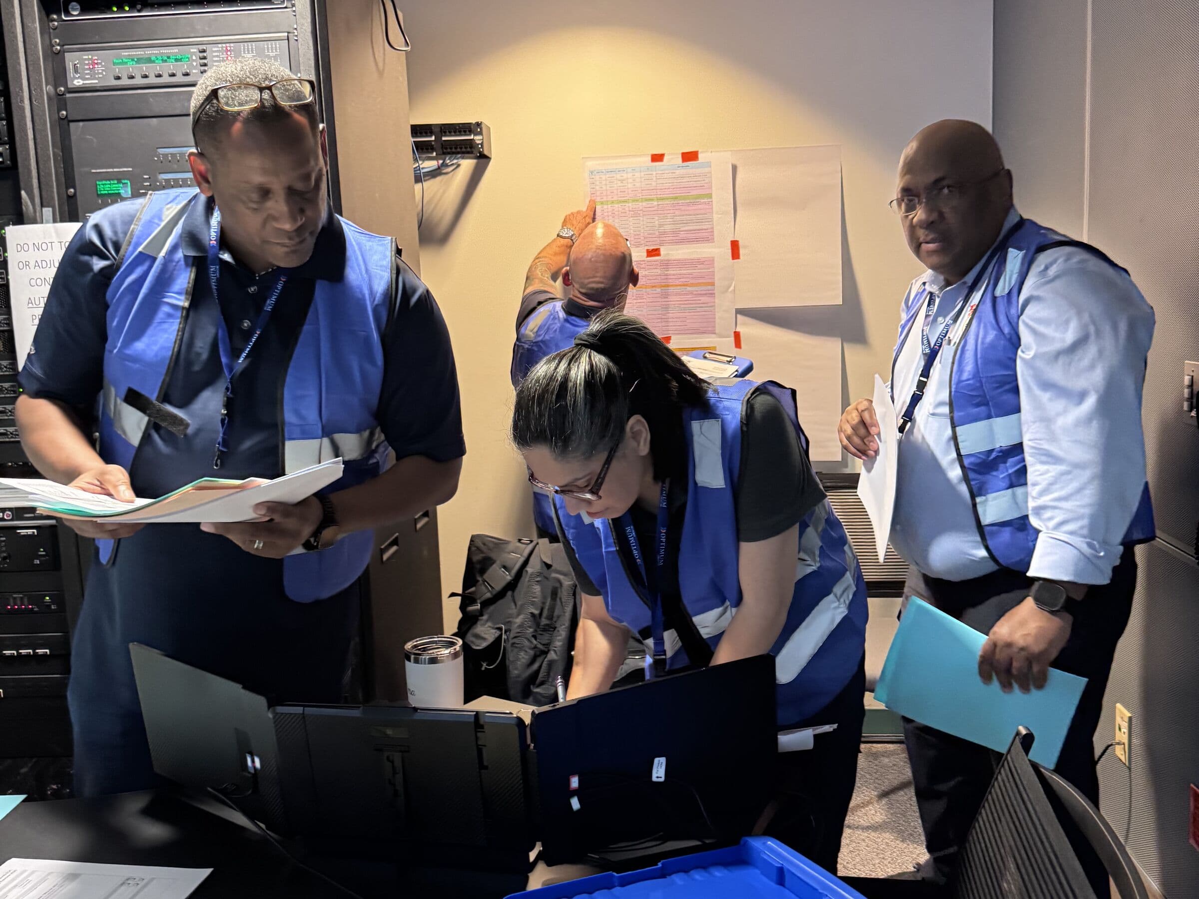 Four people in blue high-visibility vests at work in a narrow equipment room, one reading from a sheaf of papers, another bent over an open laptop, a third holding a folder, with server racks and a taped-up printed chart behind them