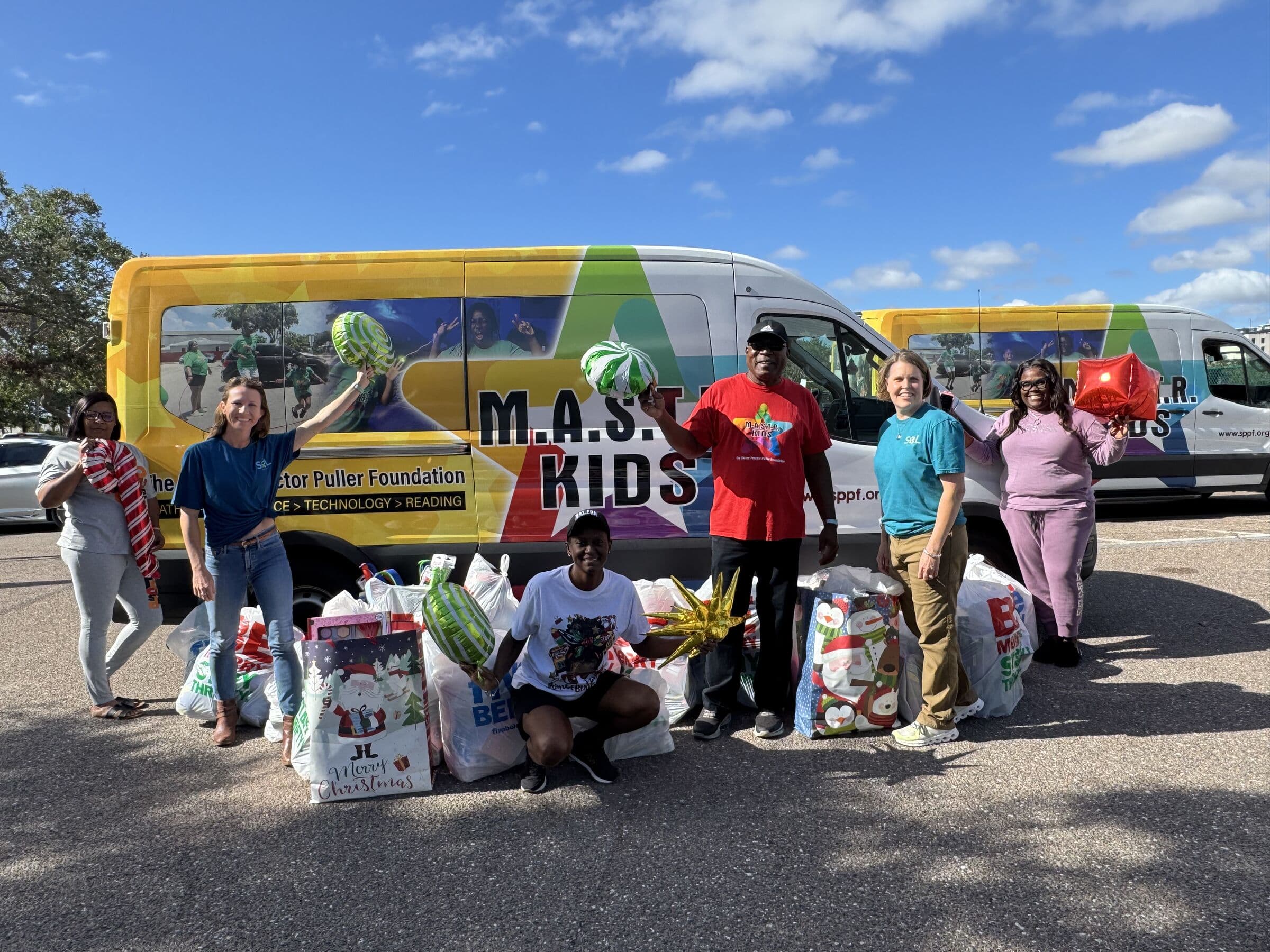 A group of people standing and crouching behind a row of filled gift bags in front of a brightly wrapped van under a blue sky