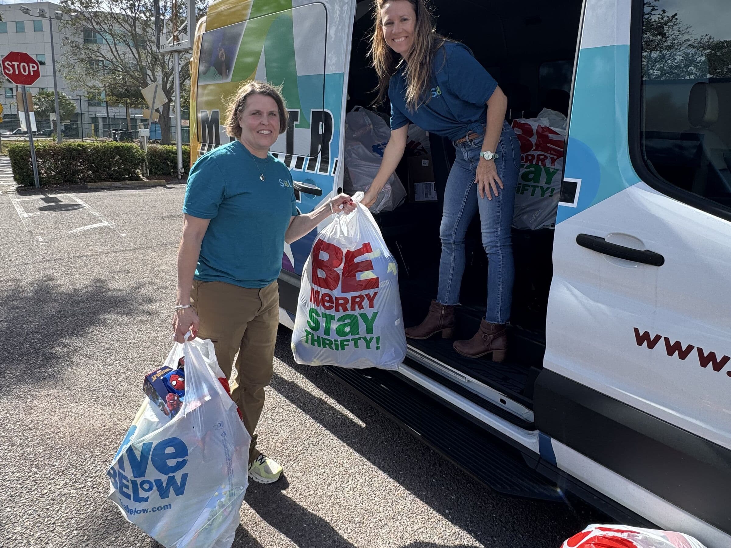 Two people carrying loaded plastic bags away from the open side door of a parked van in a car park
