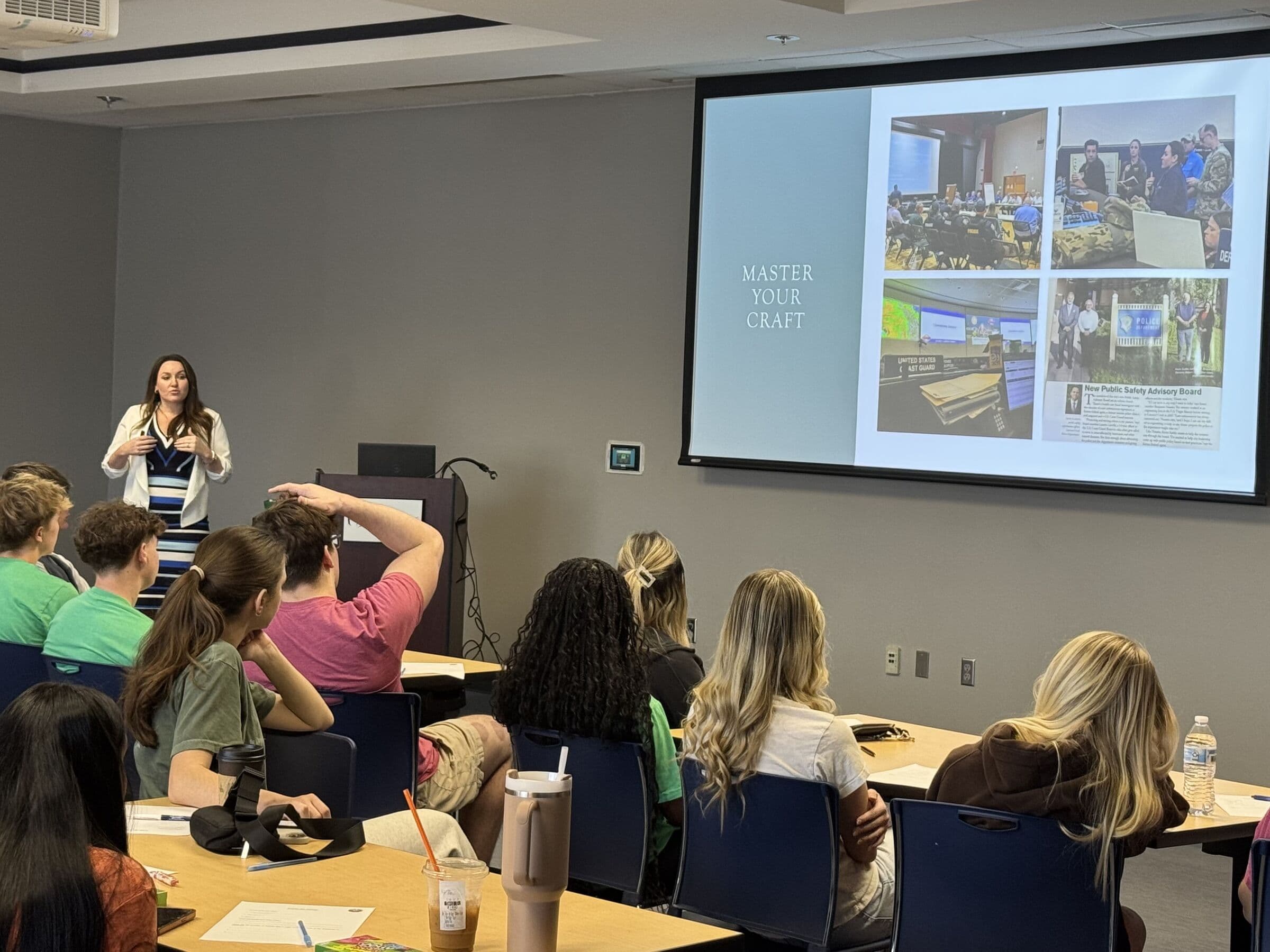 A woman presenting to a room of people seated at long tables, a projection screen of collaged photographs on the wall behind her