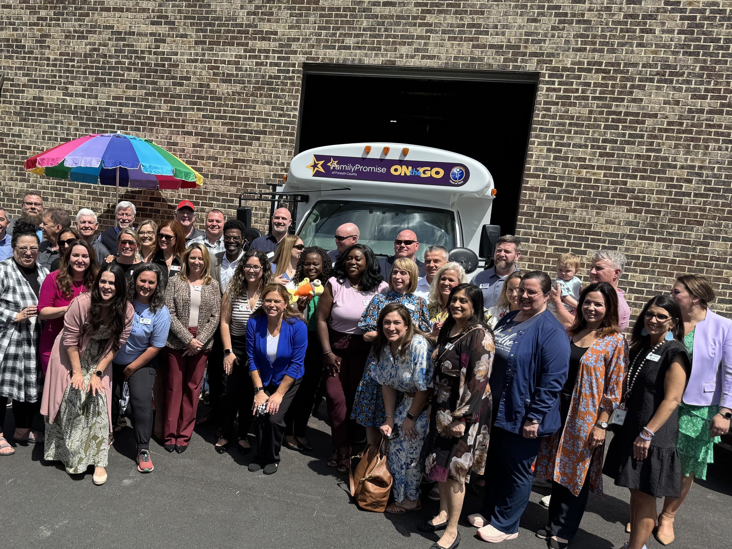A large group of people in business dress posed outside a brick building beside a white box van, one of them holding a rainbow umbrella