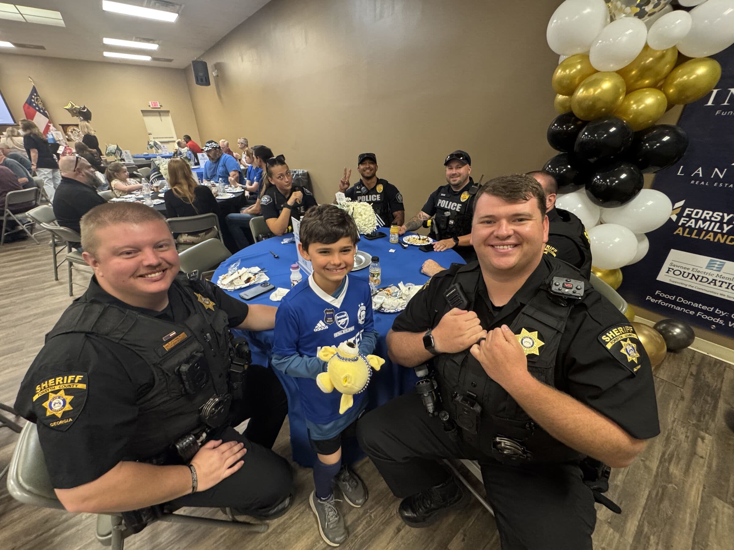 Two uniformed officers crouching either side of a boy holding a stuffed toy at a banquet table, with a black-and-gold balloon arch and people seated at tables behind them