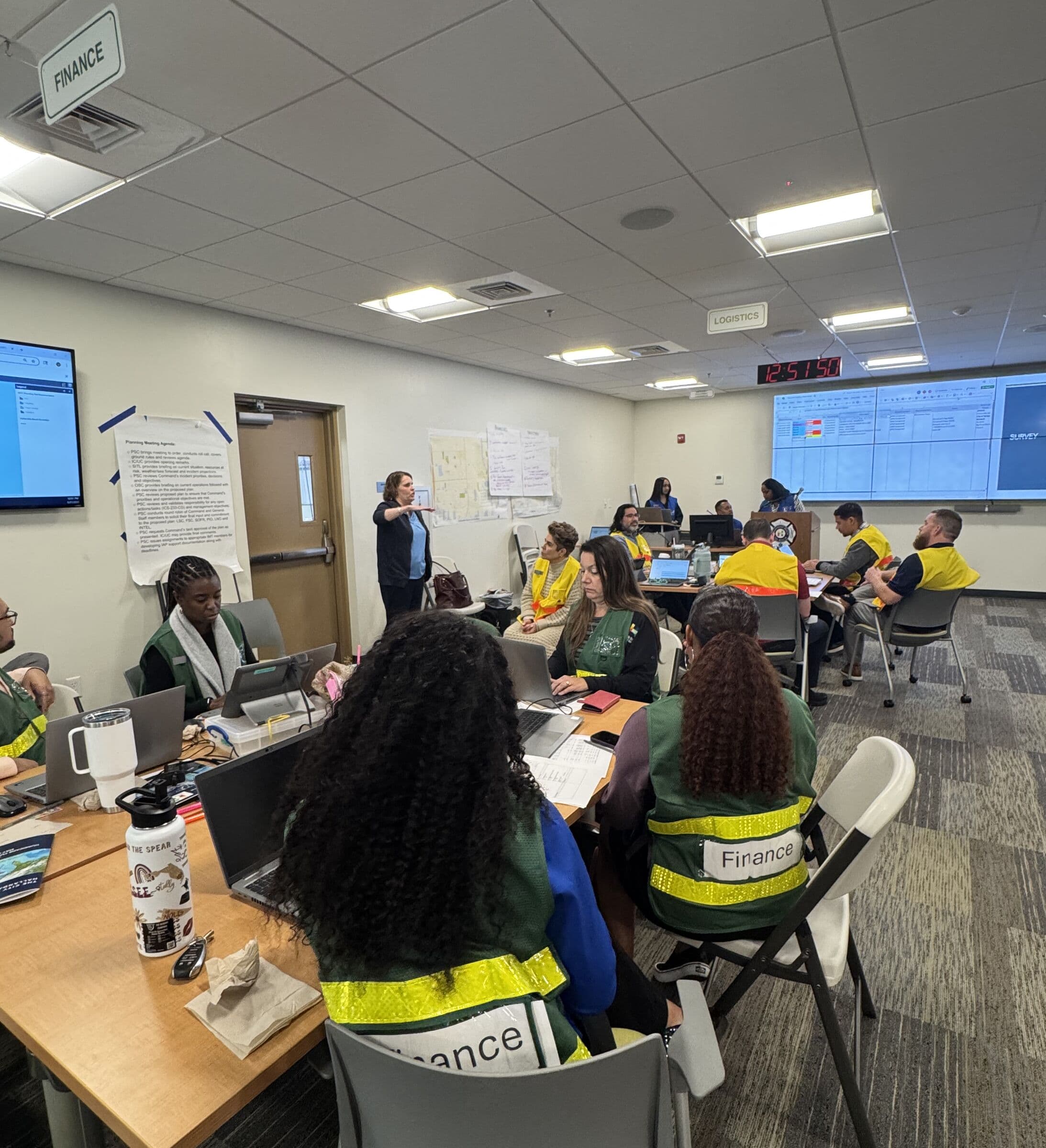 A Finance section at work during an emergency operations exercise, staff in green and yellow reflective vests at laptops around tables while a facilitator briefs the room