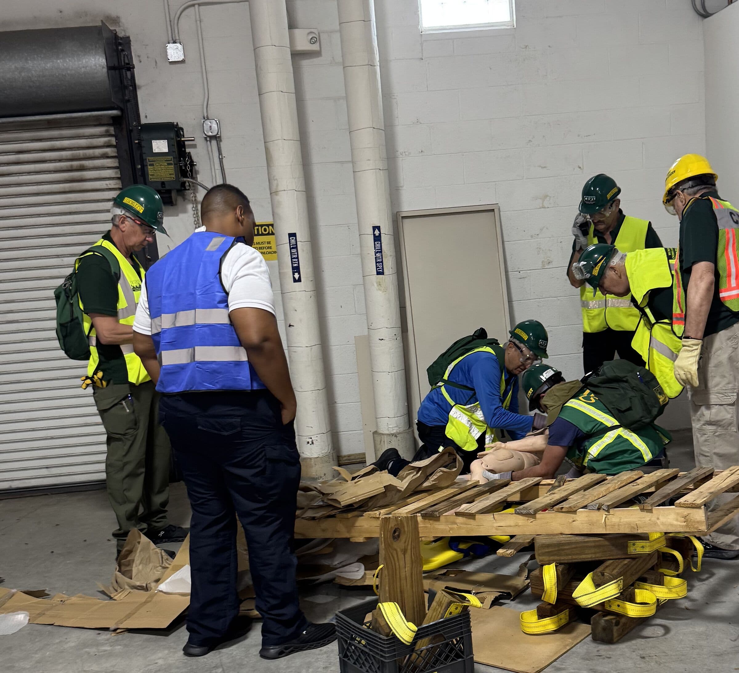 A hands-on rescue drill in a loading bay, a team in hard hats and high-visibility vests working around a training manikin under a timber pallet crib