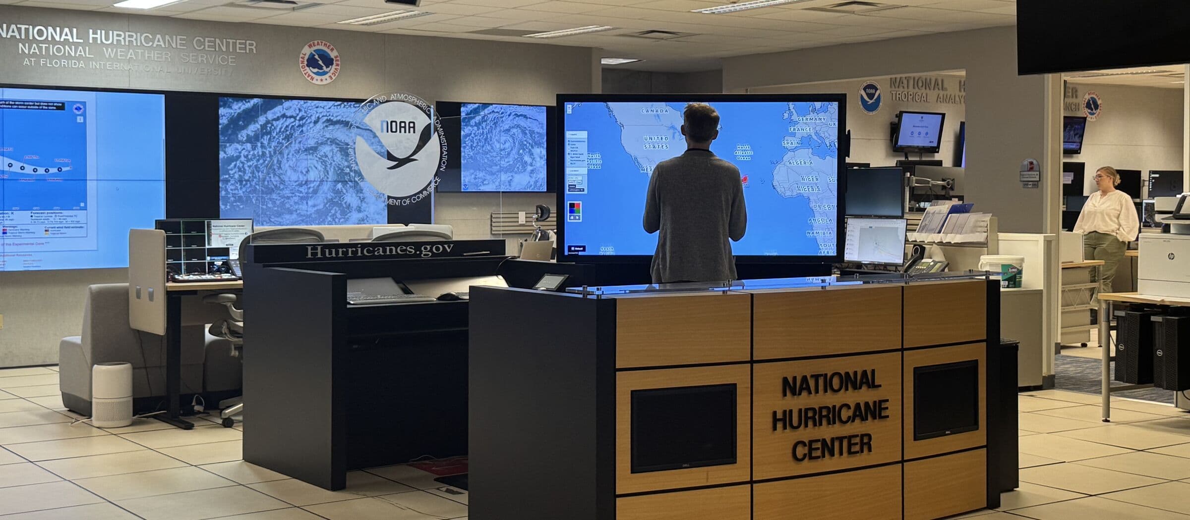 An operations floor beneath National Hurricane Center and National Weather Service lettering, with hurricane satellite imagery on a video wall and a person standing at a large world-map display