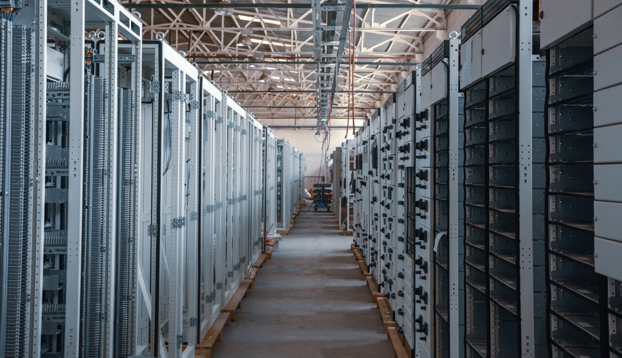 A long aisle between rows of tall grey metal equipment cabinets inside an industrial building with a steel truss roof